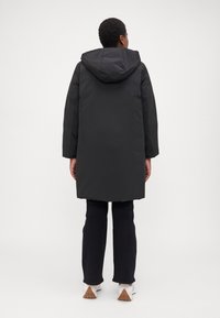 Μαύρο μακρύ μπουφάν με κουκούλα, με ίσια γραμμή, υφή επιφάνειας και πλαϊνές τσέπες, συνδυασμένο με μαύρο παντελόνι και παπούτσια.