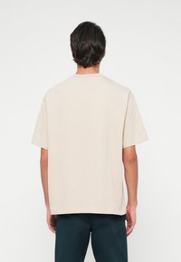 Vīrietis, tērpies īsām piedurknēm bežā T-kreklā un tumšos biksēs, stāv ar muguru pret vienkāršu balto fonu.