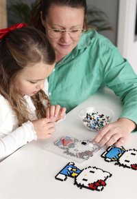 Koristetyöpakkaus eri väreissä, jossa on Hello Kitty -kuvioita muovisella naulakolla, ja vieressä kulhollinen monivärisiä helmiä.