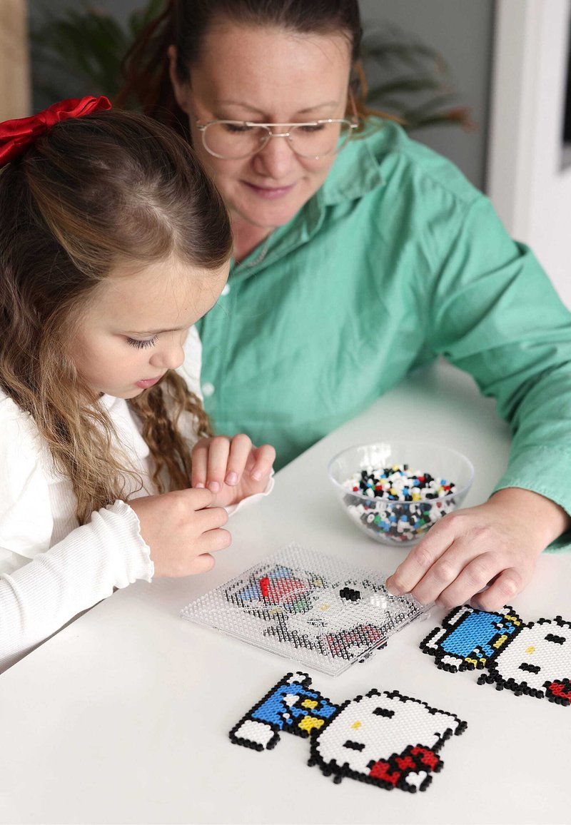 Koristetyöpakkaus eri väreissä, jossa on Hello Kitty -kuvioita muovisella naulakolla, ja vieressä kulhollinen monivärisiä helmiä.