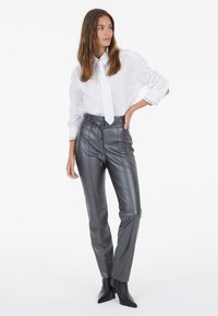 Woman wearing a white button-up shirt with a white tie, gray high-waisted leather pants, and black heeled ankle boots standing with hand on hip.