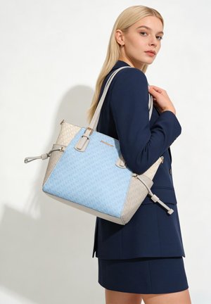 Blonde vrouw in marineblauw pak draagt een grote beige en lichtblauwe designer-totetas over haar schouder, kijkend over haar rechterschouder.