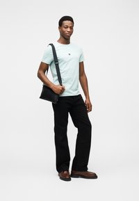 Φωτεινός γαλάζιος βαμβακερός μπλουζάκι με λογότυπο στο στήθος, μαύρο παντελόνι, καφέ δερμάτινα παπούτσια και μαύρη τσάντα ώμου. Απλό, καθημερινό ντύσιμο.