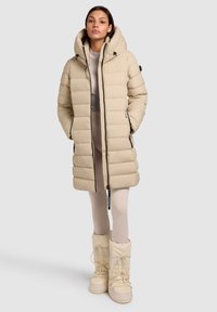 Beež värvusega puffer-jakk kapuutsiga, esikriipsuga ja küljetaskutega. Omaduseks sooniline tekstuur ja mustad detailid. Sobitatud beežide isoleeritud saapadega.