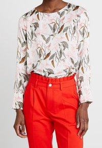 Camicetta bianca con stampa floreale in rosa, verde e marrone; abbinata a pantaloni rossi a vita alta con bottone in vita.