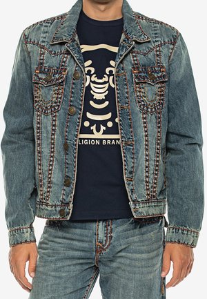 Mężczyzna w niebieskiej dżinsowej kurtce i dżinsach, granatowej koszulce z stylizowaną beżową grafiką twarzy i częściowym napisem "RELIGION BRAND".