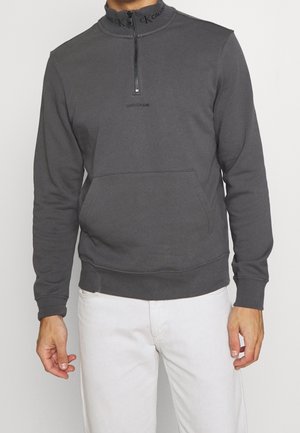 Mężczyzna ubrany w ciemnoszarą bluzę Calvin Klein z suwakiem do połowy zapinanym oraz przednią kieszenią, w białych spodniach na jednolitym tle.