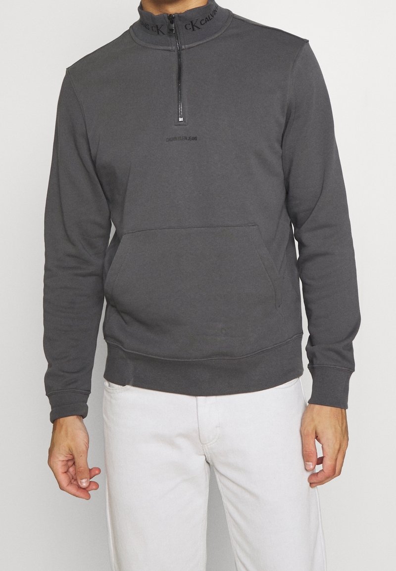 Homme portant un sweat-shirt Calvin Klein gris foncé à demi-fermeture éclair avec poche avant et pantalon blanc, sur fond uni.