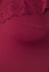Bordeaux stof met een gladde textuur en een delicate, gekartelde kantafwerking met lasergegraveerde details langs de bovenrand.