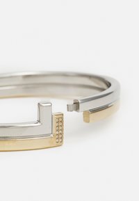 Pulsera metálica de plata y oro con forma geométrica, que presenta pequeñas piedras en la sección de oro y un diseño de cierre elegante.
