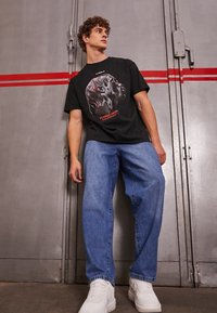 Schwarzes Grafik-T-Shirt mit einem illustrativen Design. Kombiniert mit lockeren blauen Jeans und weißen Sneakern. Hintergrund ist eine graue Wand.