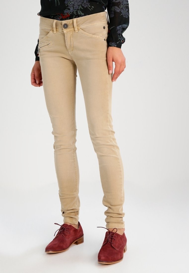 Jean skinny beige avec des accents cousus, une fermeture éclair sur la poche avant et un léger éclat. Portés avec des chaussures en suédine bourgogne à lacets.