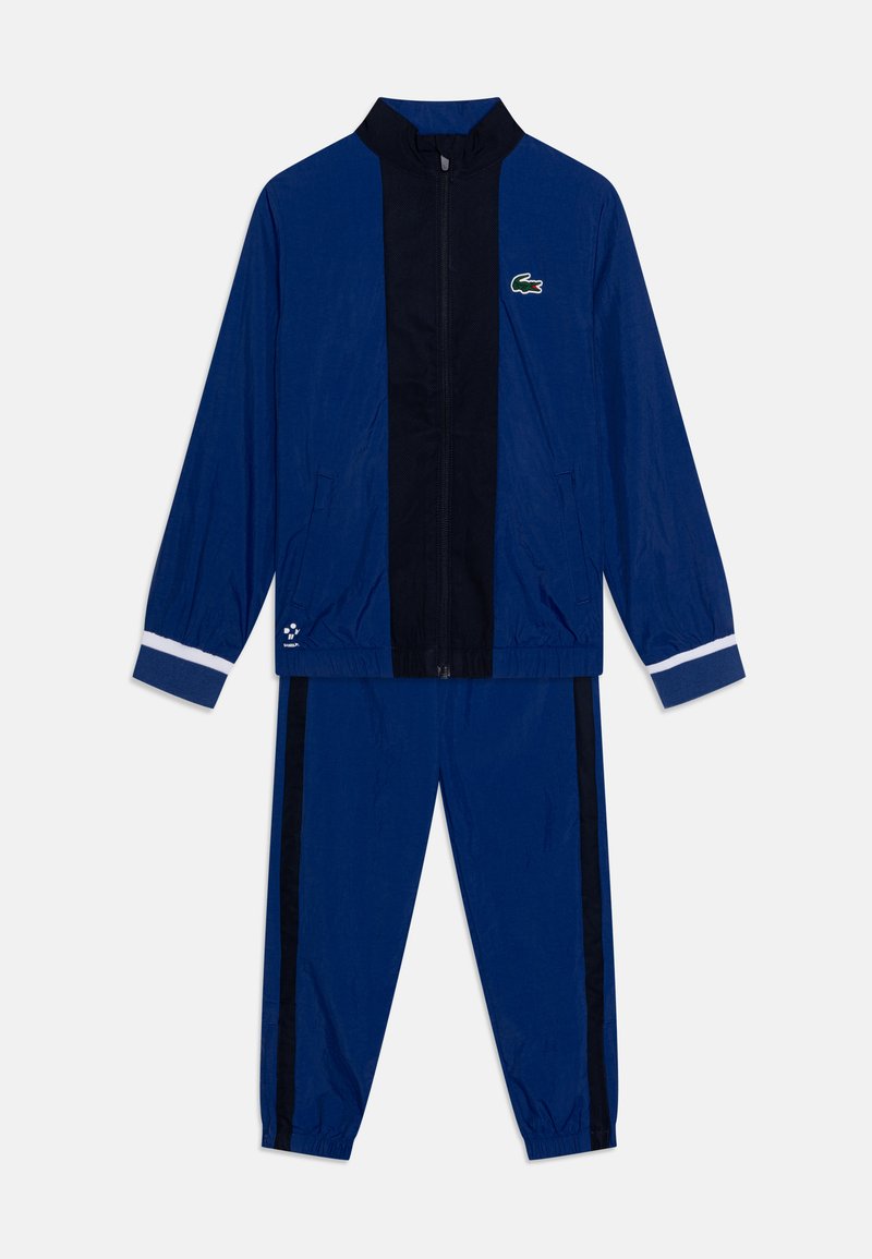 Survêtement bleu marine comprenant une veste zippée avec un col montant et des poignets rayés blancs, assorti à un pantalon fuselé avec des rayures noires sur les côtés.