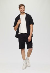 Chemise noire à manches courtes par-dessus un t-shirt blanc, associée à un short cargo noir et des baskets blanches. Cheveux de longueur moyenne et ondulés.