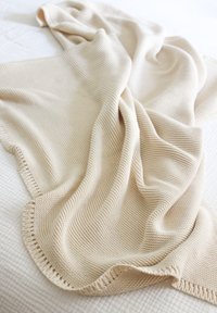 Beige knitted blanket with a textured surface, draped casually, featuring a bordered edge and soft folds against a white background.