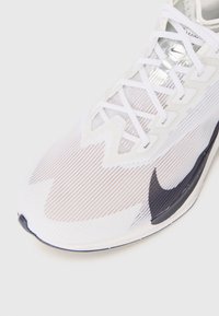 White athletic shoe with textured mesh upper, features a dark Nike swoosh, laces, and a patterned design on the toe area.