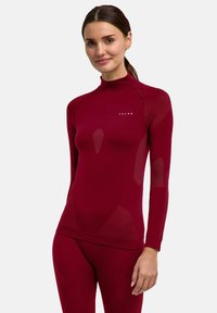 Maglia tecnica bordeaux a maniche lunghe con collo alto, caratterizzata da pannelli strutturati e design aderente. Logo visibile sul petto.