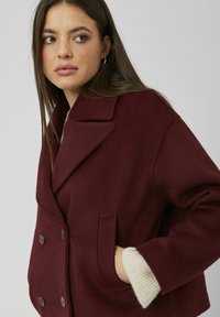 Manteau double boutonnage bordeaux avec un col cranté, en tissu de laine texturé, et poches latérales. Les poignets présentent un détail en tricot côtelé contrastant.
