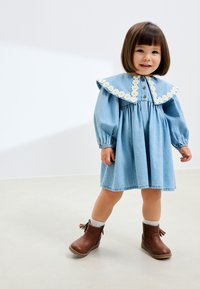 Robe en denim avec un grand col festonné orné de broderies de marguerites blanches. Manches longues bouffantes et taille froncée. Bottines marron à glands.