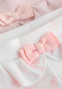 Pehmeitä vaaleanpunaisia rusetteja kiinnitettyinä herkälle valkoiselle ja vaaleanpunaiselle kuvioidulle kankaalle, jossa on teksturoidun neuloksen ja pienten silmäreikäyksityiskohtien yhdistelmä.