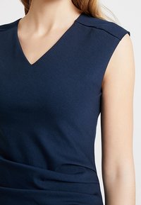 Granatowa bezrękawnik z dekoltem w serek, gładką teksturą i subtelnymi detalami na ramionach, wykonany z miękkiego, elastycznego materiału.