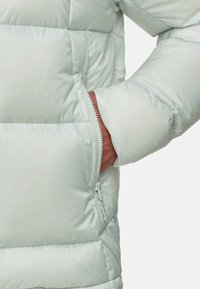 Hand gedeeltelijk in de voorste ritszak van een lichtgrijze gewatteerde winterjas met horizontale quilt.