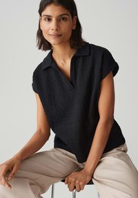 Polo shirt nera a maniche corte con un design a tessitura texturizzata. Presenta un collo a V e una vestibilità rilassata, abbinato a pantaloni beige chiaro.