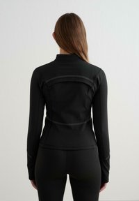 Černá sportovní bunda s přiléhavým střihem, vysokým límcem a reflexními detaily na zadní straně. Hladká textura a dlouhé rukávy.