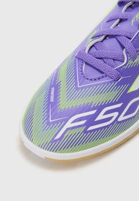 Zapato de fútbol púrpura con un patrón de chevron en acentos verdes y blancos, que presenta una parte superior texturizada y cordones para un ajuste seguro.