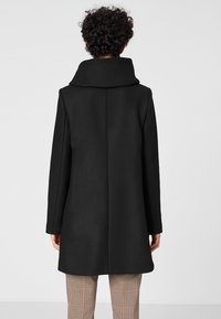 Cappotto nero di lana con colletto alto, maniche lunghe e taglio dritto. Presenta un tessuto texturizzato e un design minimale.