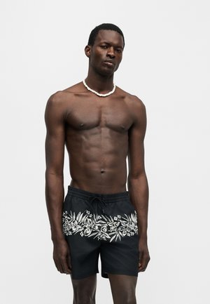 Homme musclé portant un short de bain noir avec un imprimé floral blanc et un collier de coquillages, debout devant un fond gris uni.