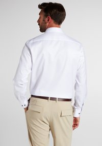 Witte langemouwen shirt met knoopmanchetten, met een recht achterpandontwerp. Gecombined met beige broek en een donkerbruine riem.
