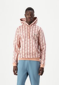 Felpa con cappuccio rosa chiaro con un motivo geometrico rosso, dotata di tasca frontale, polsini e orlo a costine. Abb binato a pantaloni della tuta blu.