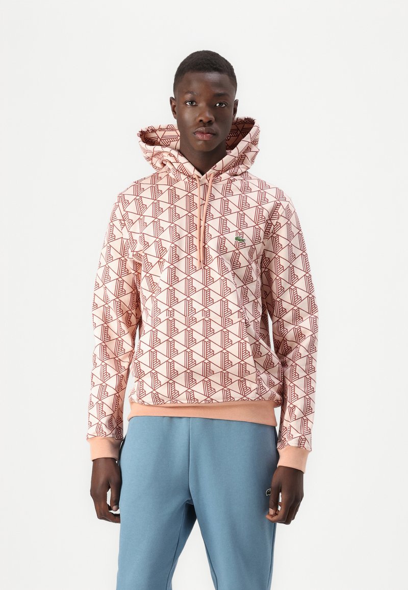 Felpa con cappuccio rosa chiaro con un motivo geometrico rosso, dotata di tasca frontale, polsini e orlo a costine. Abb binato a pantaloni della tuta blu.