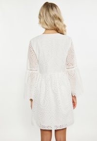 Weißes Kleid mit Broderie Anglaise Details, dreiviertel ausgestellten Ärmeln und einem gerafften Taillenbund. Der Stoff hat eine strukturierte, gemusterte Oberfläche.
