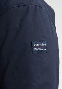 Jachetă bleu marin cu o textură în grilă. Prezintă o etichetă rectangulată cu logo-ul "Petrol Ind" pe brațul superior.