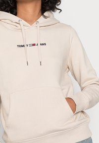 Persona che indossa un hoodie beige di Tommy Jeans con tasca frontale e lacci, mano parzialmente dentro la tasca, capelli ricci visibili.