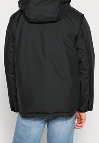 Musta eristetty takki, jossa irrotettava huppu, ribbineulepinta, painonappikiinnitys hihoissa ja väljä istuvuus.