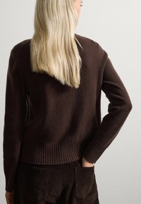 Bruine gebreide trui met geribde zoom en manchetten, met een zachte textuur en een ontspannen pasvorm. Het lange blonde haar van het model is zichtbaar.