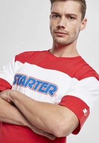 Rood-witte t-shirt met korte mouwen heeft een blauwe "STARTER" logo over de borst en een kleine steraccent op de mouw. Gemaakt van katoen.