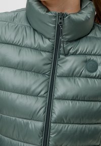 Fodrad grön jacka med hög krage och dragkedjestängning, med en matt textur, horisontell sömnad och en cirkulär logotypdetalj.