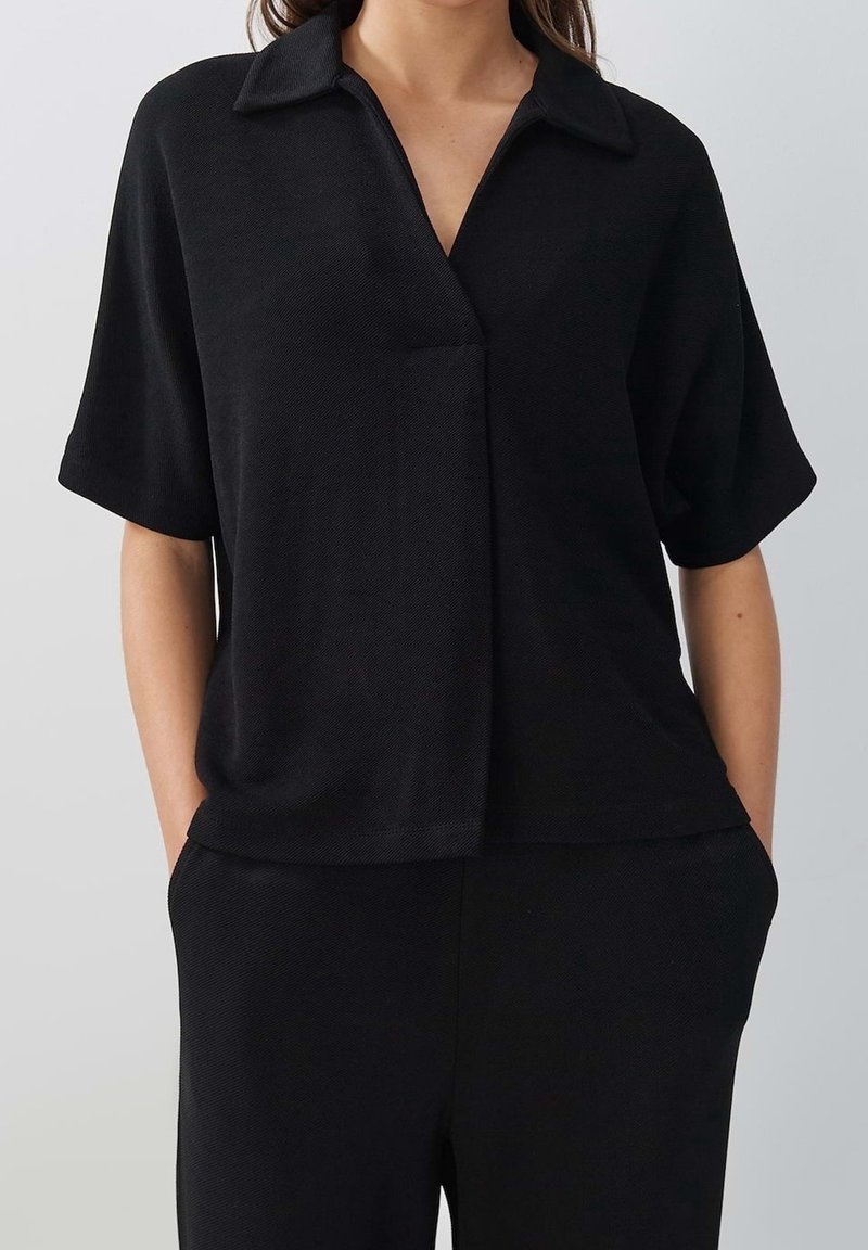 Femme portant un haut noir à manches courtes avec col et un pantalon noir assorti, les mains dans les poches, sur un fond gris clair.