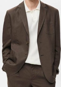 Brązowa marynarka o subtelnej fakturze, zapinana na jeden guzik jednorzędowy, z przednią kieszenią, zestawiona z białą dzianinową koszulką polo z kołnierzykiem.