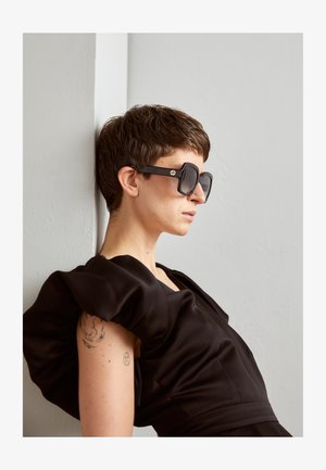 Black oversized sunglasses with a square frame and gold accents. Model wears a black puff-sleeve dress; short hair visible. Simple background.