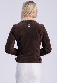 Een vrouw met lang blond haar staat met haar rug naar de kijker, gekleed in een bruine suède jas met een bontkraag en een witte gestreepte rok.