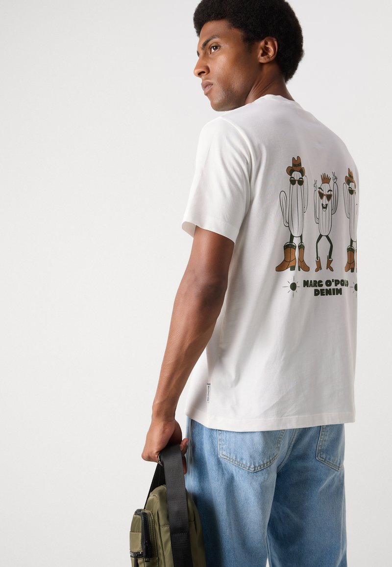 Weißes Baumwoll-T-Shirt mit verspielten Kaktusfiguren in Stiefeln und Sonnenbrillen auf dem Rücken. Das Modell hält eine grüne Tasche und trägt blaue Jeans.