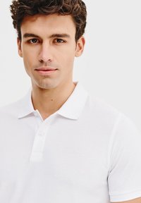 Biała koszulka polo z teksturowanej tkaniny, z dwoma guzikami przy kołnierzyku i małym logo na piersi. Krótkie rękawy i klasyczny krój kołnierza.
