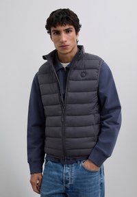 Joven vestido con un chaleco acolchado gris oscuro sobre una sudadera azul marino con cremallera y jeans de color azul claro, de pie contra un fondo claro y liso.