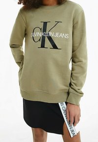 Oliwkowy sweatshirt z okrągłym dekoltem z dużym czarno-białym logo "CALVIN KLEIN JEANS". Noszony z czarną spódnicą i białym detalem metki.