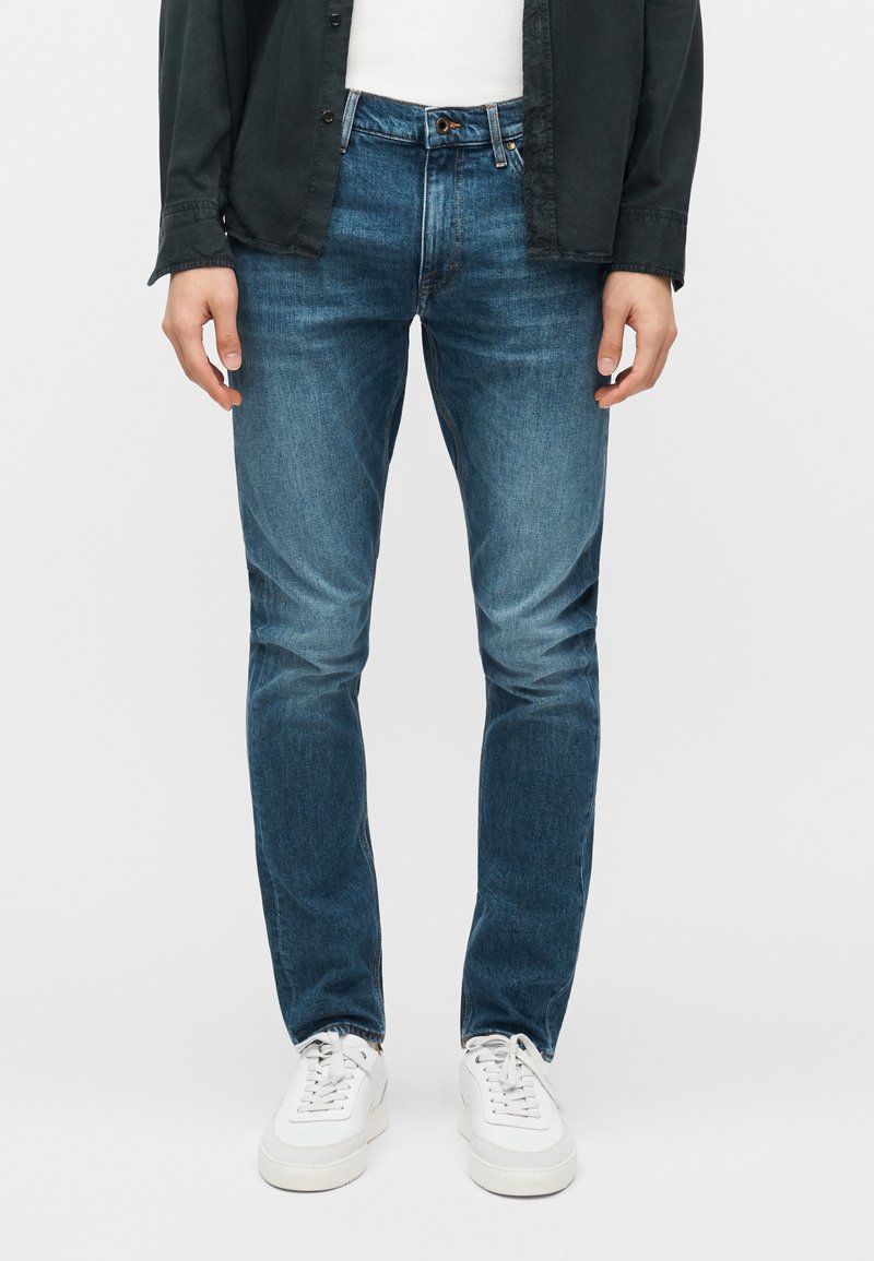 Άνδρας που φοράει στενά τζιν μέτριας μπλε απόχρωσης, λευκά αθλητικά παπούτσια, λευκό πουκάμισο και μαύρο ανοιχτό πουκάμισο με κουμπιά, σε απλό φόντο.